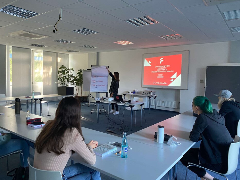 Mehrere Personen sitzen in einem Seminarraum, vorne steht eine Präsentation mit FLYERALARM-Logo auf der Leinwand.